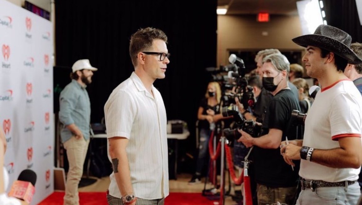 Ricky in a black cowboy hat interviewing Bobby Bones on the iHeartCountry Festival carpet, a bank of press cameras behind them.